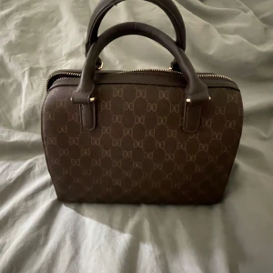 Brun handväska från Don Donna - Snygg brun handväska från Don Donna med klassiskt monogrammönster och guldfärgade detaljer. Väskan har två handtag, avtagbar axelrem och dragkedja upptill. Tillverkad i syntetmaterial med rymligt fack och innerficka med dragkedja. Kan alltid diskutera pris. Skriv för frågor och bilder🥰🥰