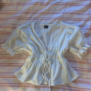 Vit blus med snörning från Gina Tricot - Supersöt vit blus från Gina Tricot i storlek XXS. Blusen har korta puffärmar, v-ringning och snygg snörning i midjan som ger en feminin siluett. Tillverkad i ett lätt, strukturerat tyg som känns luftigt och fräscht. Perfekt för vår och sommar!