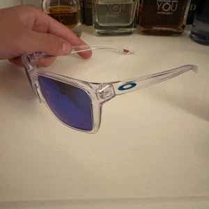 Oakley solglasögon med blå spegelglas - Snygga Oakley solglasögon med transparent båge och blå spegelglas. Klassisk fyrkantig form och cool Oakley-logga på sidan. Perfekta för dig som vill sticka ut med en modern och sportig look.