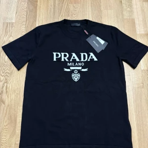 Svart t-shirt från Prada med logga - Svart t-shirt från Prada med vit broderad logga och texten 'PRADA MILANO' på bröstet. Klassisk rund halsringning och korta ärmar. T-shirten är i mjuk bomull och har en clean, stilren look som passar perfekt till jeans eller shorts.