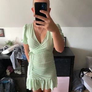 Grön rutig omlottklänning Gina Tricot - Supersöt kort grön och vit rutig omlottklänning från Gina Tricot. Klänningen har v-ringning, puffiga korta ärmar, knyt i midjan och volangkant nertill. Perfekt för sommarens alla tillfällen!
