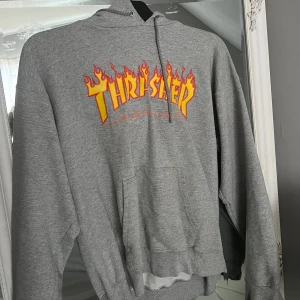 Grå Thrasher hoodie med flames - Säljer en grå hoodie från Thrasher med ikoniskt flames-tryck i gult och rött på bröstet. Klassisk känguruficka, huva med snörning och ribbade muddar. Perfekt för dig som gillar streetwear och skate-stil.