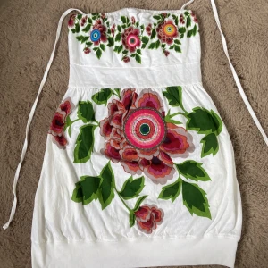 Vit blommig kortklänning Desigual M - Superfin vit kortklänning från Desigual med färgglada blommor och gröna blad, samt en grön glittrig fjäril på baksidan. Klänningen har axelbandslös modell med knytband och är perfekt för sommarens alla tillfällen.🌸