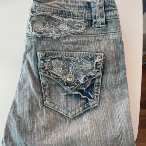Ljusblå jeans med spetsdetaljer - Snygga ljusblå jeans med raka ben och unika spetsdetaljer på bakfickorna och ovanför fickorna. Jeansen har slitna kanter och dekorativa knappar med bling. Perfekt för dig som vill sticka ut med coola detaljer och en avslappnad vibe.