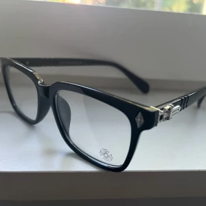 Svarta glasögon med silverdetaljer Chrome Hearts - Stilrena svarta glasögon från Versace med markerad båge och fyrkantig form. Snygga silverdetaljer vid gångjärnen och diskret logga på skalmen. Perfekta för dig som vill ha en klassisk men ändå trendig look.