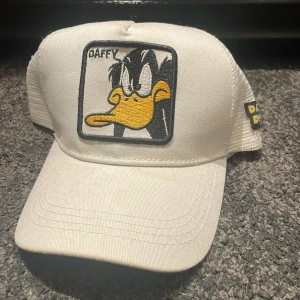 Vit Daffy Duck keps med mesh - Cool vit keps med broderad Daffy Duck på framsidan och mesh på sidorna för extra ventilation. Kepsen har böjd skärm och justerbar passform. Daffy Duck-logga även på sidan. Perfekt för dig som gillar Looney Tunes och vill sticka ut.