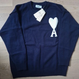 Ami Paris tröja - Snygg mörkblå stickad tröja från Ami Alexandre Mattiussi med stor vit hjärtlogga och bokstaven A på bröstet. Klassisk rund halsringning och ribbade muddar. Perfekt för dig som gillar stilrena och trendiga plagg.