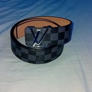 rutigt skärp med spänne - Snyggt bälte i svart och grått med klassiskt rutigt mönster och stort spänne i silver. Bältet är gjort i skinn och har en exklusiv känsla. Perfekt accessoar för att lyfta din outfit med en lyxig touch.