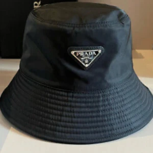 Svart bucket hat från Prada - Stilren svart bucket hat från Prada med ikonisk triangellogo framtill i silver. Hatten är tillverkad i glansig syntet och har klassisk form med bred, nedåtlutande brätte. Perfekt accessoar för att lyfta din outfit med en lyxig touch.