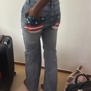 Blå jeans med USA-flagga på bakfickor( low waist)  - Säljer ett par blå jeans med coola bakfickor som har USA-flagga med stjärnor och ränder. Jeansen har hög midja och bootcut-ben, vilket ger en snygg retrovibe. Tillverkade i klassiskt denimtyg och passar perfekt för dig som vill sticka ut.