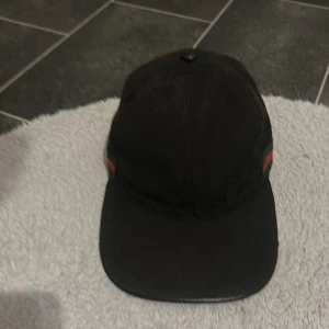 Svart keps med röd-gröna detaljer - Svart keps med böjd skärm och diskreta röd-gröna ränder på sidorna. Klassisk modell med knapp på toppen och paneler. Perfekt accessoar för en clean streetstyle-look. Mer info kom privat!!!