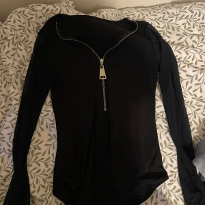 Svart långärmad topp med dragkedja - Säljer en svart långärmad topp med en snygg silverfärgad dragkedja framtill. Toppens material är stretchigt och figurnära, perfekt för att skapa en cool och stilren look. Passar dig som gillar enkla men trendiga plagg.