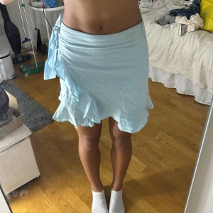 Ljusblå omlottkjol med volang SHEIN XS - Supersöt ljusblå kortkjol från SHEIN i omlottmodell med knyt i sidan och volang längs nederkanten. Kjolen är i ett tunt, lätt material som passar perfekt till varmare dagar. Enkel att styla med crop top eller t-shirt för en chill look.