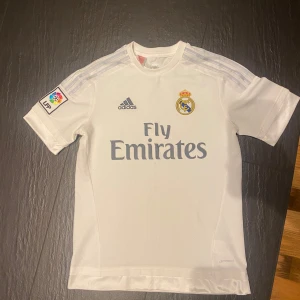 Real Madrid Adidas fotbollströja vit - Snygg vit Real Madrid fotbollströja från Adidas med klubbmärke och Fly Emirates-tryck på bröstet. Tröjan har korta ärmar, La Liga-märke på ena ärmen och är tillverkad i lätt och ventilerande material. Perfekt för dig som älskar fotboll och vill bära klubbens färger.