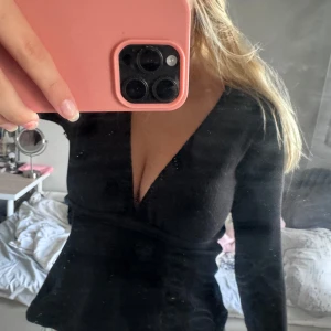 Svart omlottop med v-ringning - Säljer en svart omlottop med  v-ringning och långa ärmar. Toppen har knyt i sidan vilket ger en snygg passform och markerad midja. Perfekt för dig som gillar stilrena och trendiga plagg med lite edge.