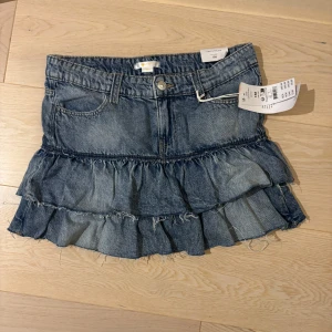 Jeanskjol med volanger från Gina Tricot - Supersöt jeanskjol från Gina Tricot i klassisk blå denim. Kjolen har två lager volanger med råa kanter för en cool och trendig look. Klassisk femficksmodell med knapp och dragkedja framtill. Perfekt för dig som gillar en avslappnad men ändå stylish vibe.