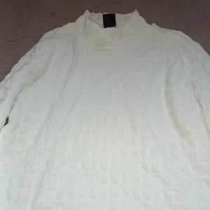 Vit långärmad topp - Säljer en vit långärmad topp med en unik textur. Toppen har en hög krage och ett subtilt rutmönster som ger en stilren look. Perfekt för att lägga till en touch av elegans i din garderob.