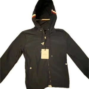 Moncler windbreaker/vindjacka Marinblå - Snygg Marinblå vindjacka från Moncler med huva och dragkedja. Jackan har långa ärmar och en stilren design med röd och vit randdetalj på huvan. Perfekt för blåsiga dagar. Helt ny jacka har endast tagit ut den för o ta bild till annonsen. Priset kan diskuteras! finns det några frågor så är det bara att ställa dem! Färgen ser mer svart ut på bilden men är marinblå om det önskas fler bilder är det bara att skriva! Nfc tag finns.