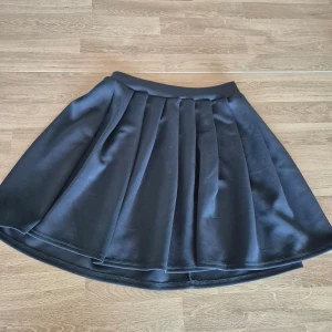 Svart kjol med veck - Snygg svart kjol med veck och dragkedja i ryggen. Elastisk midja. Tillverkad i en blandning av polyester och bomull för en skön passform. Perfekt för olika tillfällen!