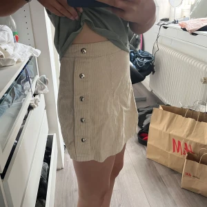 Beige kortkjol med knappar - Säljer en stilren beige kortkjol med knappar framtill. Kjolen har en dragkedja på sidan och är tillverkad i ett ribbat material. Perfekt för en trendig look!