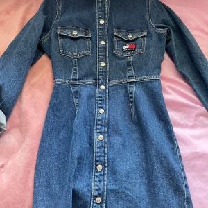 Jeansklänning från Tommy Jeans - Snygg jeansklänning från Tommy Jeans i en klassisk blå denim. Klänningen har långa ärmar och knappar hela vägen ner. Två bröstfickor med knapp och en broderad logga på ena fickan ger en cool touch.