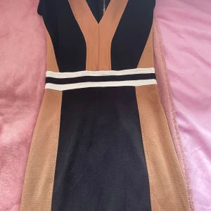 Svart och beige klänning från H&M - Snygg svart och beige klänning från H&M med en v-ringad design och dragkedja i ryggen. Klänningen har en kontrasterande vit rand i midjan som ger en stilren look. Perfekt för en chic och elegant stil.