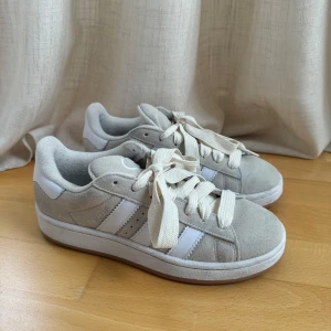 Beige sneakers från Adidas - Snygga beige sneakers från Adidas med vita detaljer och snörning. Skorna har en klassisk design med tre ränder på sidan och en bekväm sula. Perfekta för en stilren vardagslook. Dock har de fått lita avfrågning av jeans men annars använts cirka 3 gånger sammanlagt