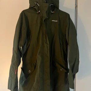 Grön parkas från Didriksons - Säljer en grön parkas från Didriksons med huva och dragsko. Jackan har långa ärmar och stängs med dragkedja och knappar. Perfekt som allvädersjacka