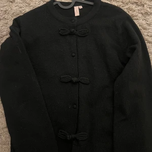 Svart kofta från Nelly - Säljer en stilren svart kofta från Nelly med dekorativa knytningar framtill. Koftan har långa ärmar och en rund halsringning, perfekt för en elegant look.