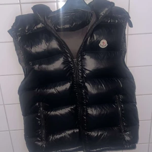 Svart dunväst från Moncler - Snygg svart dunväst från Moncler med glansig finish och dragkedja framtill. Västen har en hög krage och Moncler-logga på bröstet. Perfekt för kyliga dagar när du vill hålla stilen.