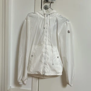 Vit vindjacka från Moncler - Säljer en stilren vit vindjacka från Moncler med huva och dragkedja. Jackan har långa ärmar och en broderad logotyp på ärmen. Perfekt för vår och sommar med sin lätta design och snygga detaljer. Skick 8/10 storlek 2 som motsvarar M  