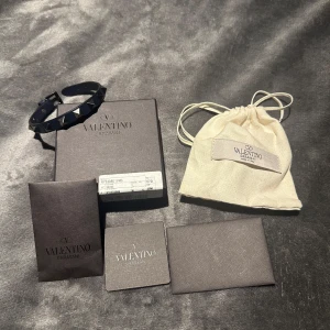 Mörkblått armband med nitar från Valentino - Säljer ett stilrent svart armband från Valentino med pyramidformade nitar. Armbandet har justerbar passform och kommer med originalförpackning och påse. Perfekt för att ge en edgy touch till din outfit. Armbandet är köpt utomlands på en Valentino butik som en present men aldrig använt då jag inte gillar armband.