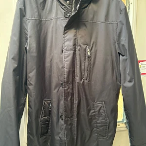 Svart rock från Matinique - Snygg svart rock från Matinique med dragkedja och knappar. Jackan har en quiltad insida och flera praktiska fickor. Perfekt för kyligare dagar med sin långa design och stilrena look. Löstagbart innerfoder. Sparsamt använd. 