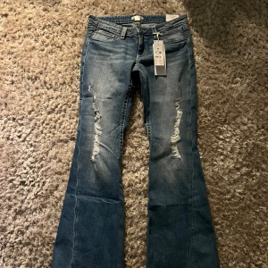 Blå lowrise bootcut jeans med slitningar - Snygga blå lowrise  bootcut jeans med slitningar på framsidan. Jeansen har en klassisk femficksdesign och dekorativa bakfickor med knappar. Perfekta för en avslappnad stil. Midjemått ca 37cm och Innerbenslängd ca 75-76cm.