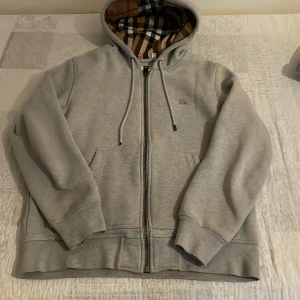 Grå hoodie med mönstrat foder - Säljer en stilren grå hoodie från Burberry med dragkedja och mönstrat foder i klassiskt rutigt mönster. Tröjan har en broderad logga på bröstet och justerbar huva med snören. Perfekt för en avslappnad och trendig look.
