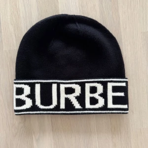 Burberry mössa svart  - Snygg svart mössa från Burberry med vit text. Perfekt för att hålla värmen med stil. Den har en bred uppvikt kant med märkets namn i kontrasterande vitt. Mössan är aldrig använd och är helt ny som ni ser på bilderna! Tagsen till mössan är också kvar! 