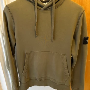 Olivgrön hoodie från Stone Island - Säljer en snygg olivgrön hoodie från Stone Island med klassisk logga på ärmen. Tröjan har en känguruficka framtill och justerbar huva med dragsko. Perfekt för en avslappnad stil. Pris kan diskuteras vid en snabb affär.