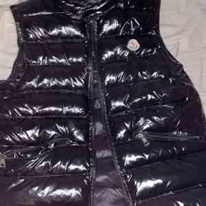 Svart glansig dunväst från Moncler - Snygg svart glansig dunväst från Moncler med dragkedja och två praktiska fickor framtill. Perfekt för att hålla värmen med stil. Moncler-logga på bröstet ger en exklusiv touch. Dragkedjan är sönder