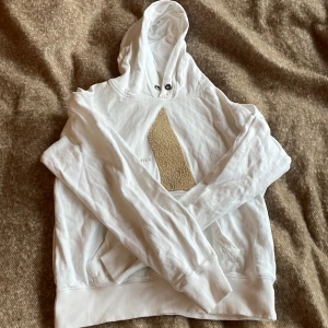 Vit hoodie med beige detalj från Milk - Här har vi en mysig och gosig vit Milk Atelier. Har använts några gånger och har ett bra skick för det. Hoodien har dock en pytteliten fläck på ryggen, men den syns inte så tydligt. 
