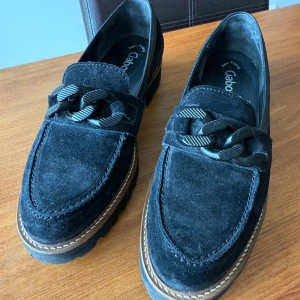 Svarta mockaloafers från Gabor - Snygga svarta loafers i mocka från Gabor med dekorativ kedja på ovansidan. Skorna har en robust sula med bra grepp och en stilren design som passar perfekt för både vardag och mer uppklädda tillfällen. Säljes på grund av storleken. Känns som en stor 39:a. 