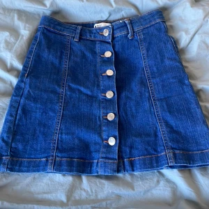 Blå jeanskjol från Gina - Snygg blå jeanskjol från Gina  med knappar framtill. Kjolen har en klassisk design med bälteshällor och en A-linjeformad passform. Perfekt för en avslappnad stil.