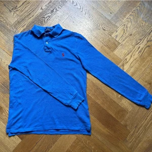 Blå pikétröja från Ralph Lauren - Säljer en snygg blå pikétröja från Ralph Lauren med röd broderad logga på bröstet. Tröjan har långa ärmar och en klassisk krage med knappar. Perfekt för en stilren look!