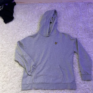 Grå hoodie från Lyle & Scott - Säljer en stilren grå hoodie från Lyle & Scott med deras ikoniska logga på bröstet. Tröjan är tillverkad i 100% ekologisk bomull och har en klassisk passform med långa ärmar och en bekväm huva. Perfekt för en avslappnad look!