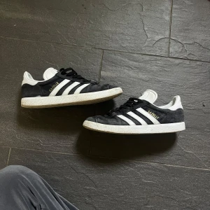 Adidas Gazelle marinblå sneakers mocka - Klassiska Adidas Gazelle sneakers i marinblå mocka med vita detaljer och de ikoniska tre ränderna på sidorna. Skorna har vit plös och häl med Adidas-logga, samt svart snörning och platt vit sula. Perfekta för en clean och sportig stil.