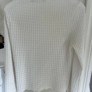 Vit långärmad stickad tröja - Vit långärmad stickad tröja med vågigt strukturmönster över hela plagget. Tröjan har en klassisk rak passform och är perfekt för lager-på-lager. Enkel och stilren design som passar till det mesta.