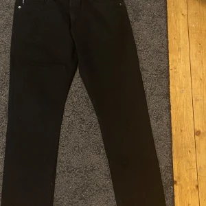 Svarta jeans från Emporio Armani - Snygga svarta jeans från Emporio Armani med klassisk rak passform och diskret logga i silver på bakfickan. Byxorna har svarta sömmar med en cool röd detalj på insidan och läderpatch med logga bak i midjan. Perfekt för dig som gillar stilrena designer.