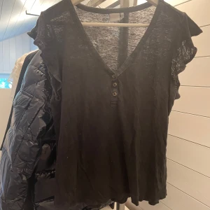 Svart linnetopp med volangärm H&M - Svart linnetopp från H&M i svalt linne. Toppen har v-ringning med tre knappar framtill och korta volangärmar som ger en chill vibe. Perfekt för varma dagar när du vill ha något enkelt men ändå snyggt.