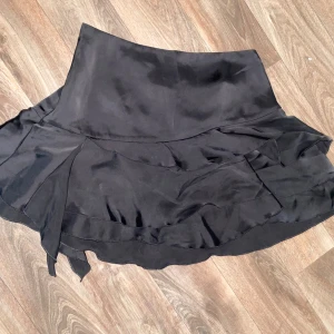 Svart omlottkjol med volang - Snygg svart kortkjol i omlottmodell med volangdetaljer. Kjolen har en lätt glansig yta och knytband i sidan för en cool och feminin look. Perfekt för dig som gillar trendiga och stilrena plagg.