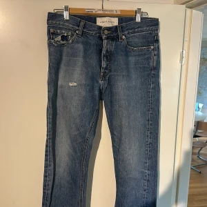 Blå jeans från A Day's March, strl 29 - Säljer ett par klassiska blå jeans från A Day's March, modell Denim No.2. Jeansen har rak passform, fem fickor och snygga slitningar vid fickan och på låret. Tillverkade i robust denim med en tidlös look.
