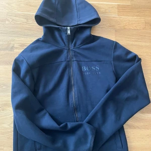 Marinblå hoodie från Hugo Boss - Snygg marinblå hoodie från Hugo Boss med dragkedja framtill och diskret logga på bröstet. Tröjan har huva, långa ärmar och är tillverkad i mjuk bomull. Perfekt för en chill och stilren look.